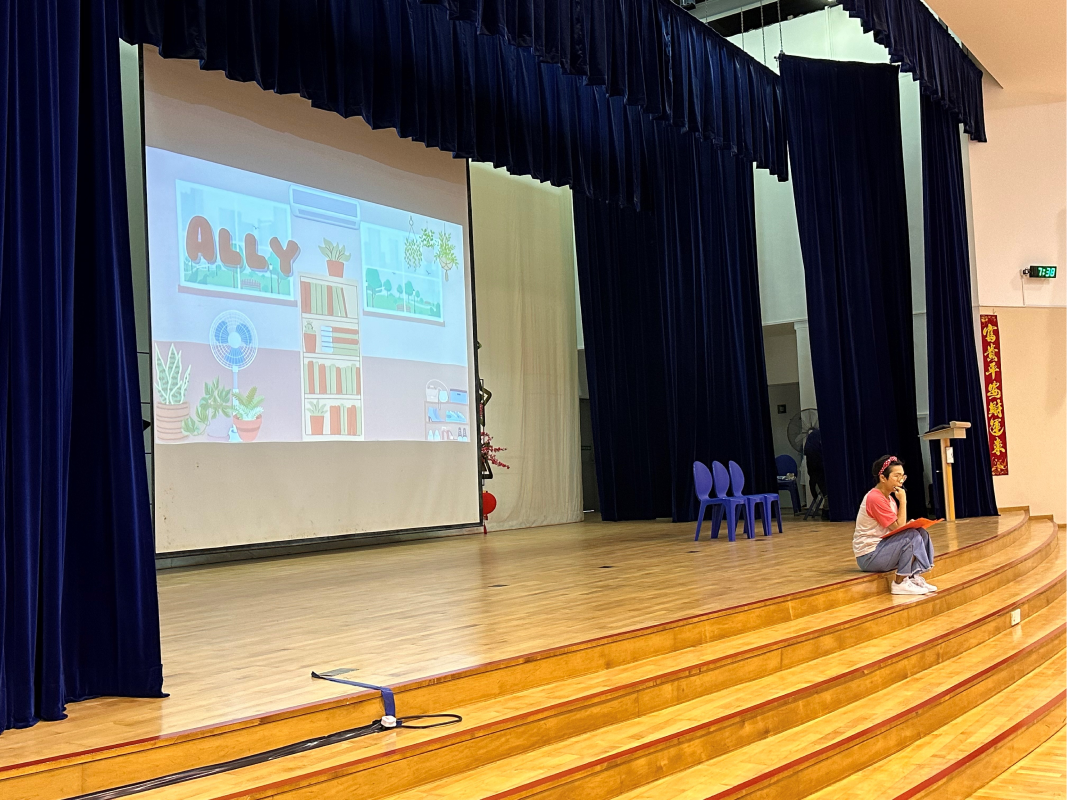 A person sits on stage steps beside a projected screen showing a cartoon titled "ALLY" in a school auditorium.