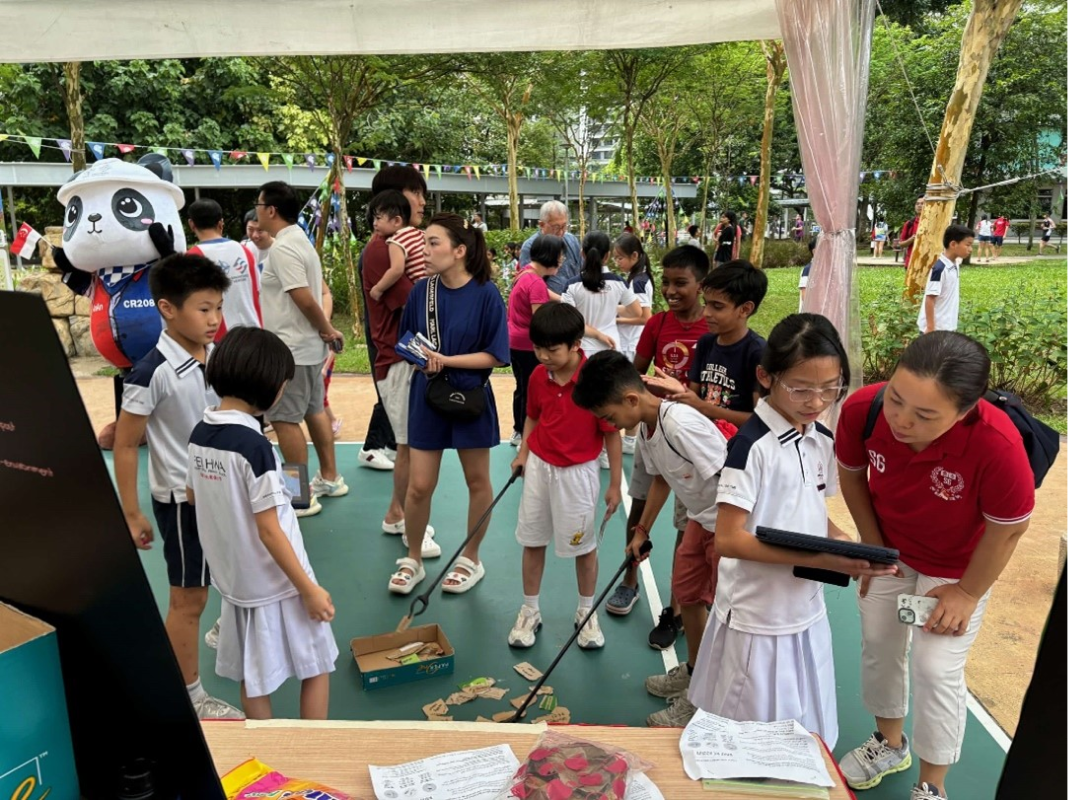 Children in school uniforms participate in an outdoor activity booth with adults, colorful bunting, and a panda mascot balloon.