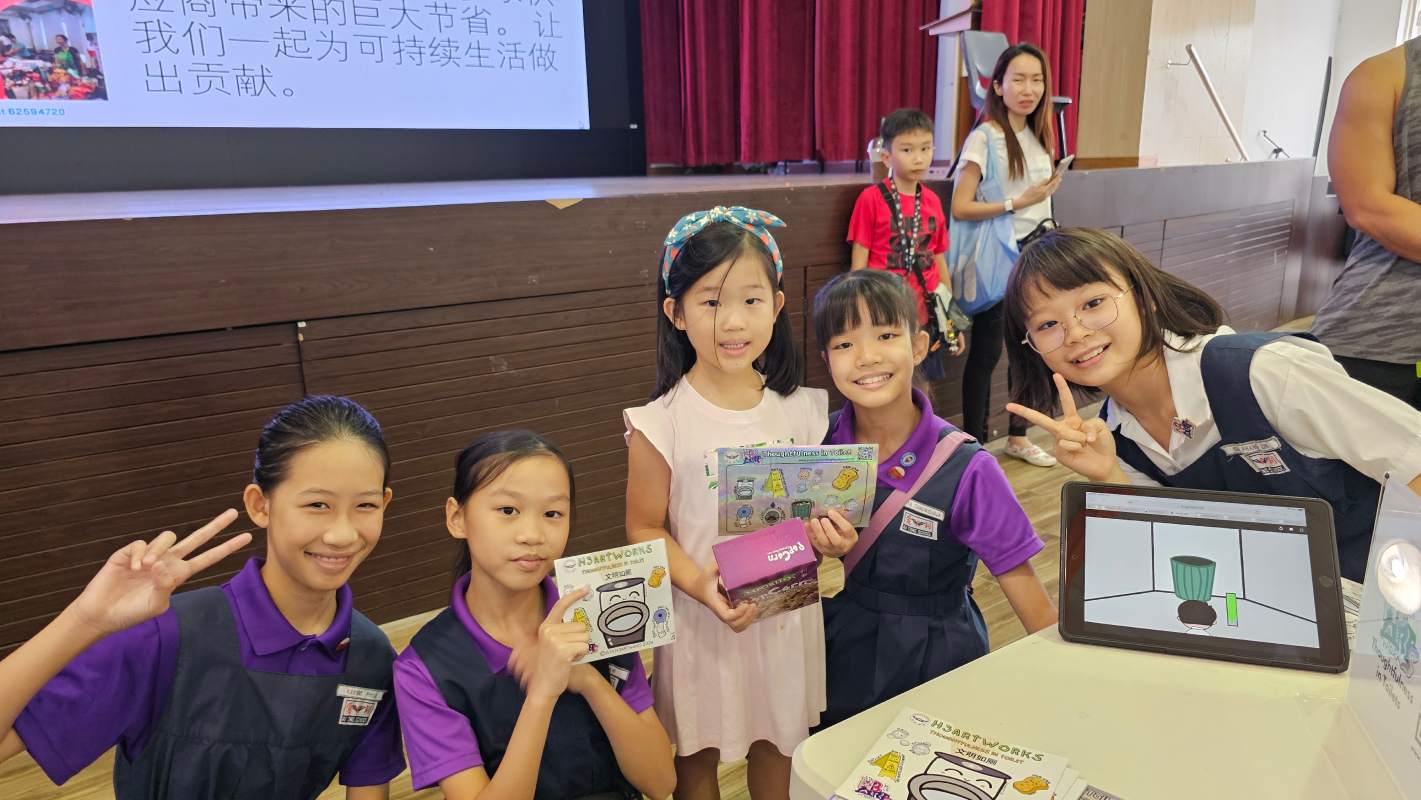 Five smiling girls pose together holding cards and a box near a tablet displaying a green bin on screen.