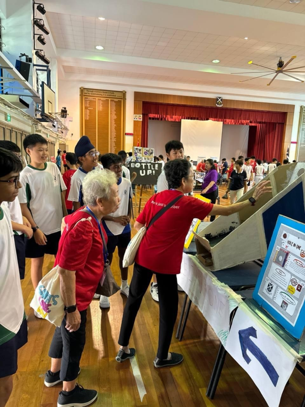 Students and adults gather around project displays in a school hall during what appears to be a science fair.