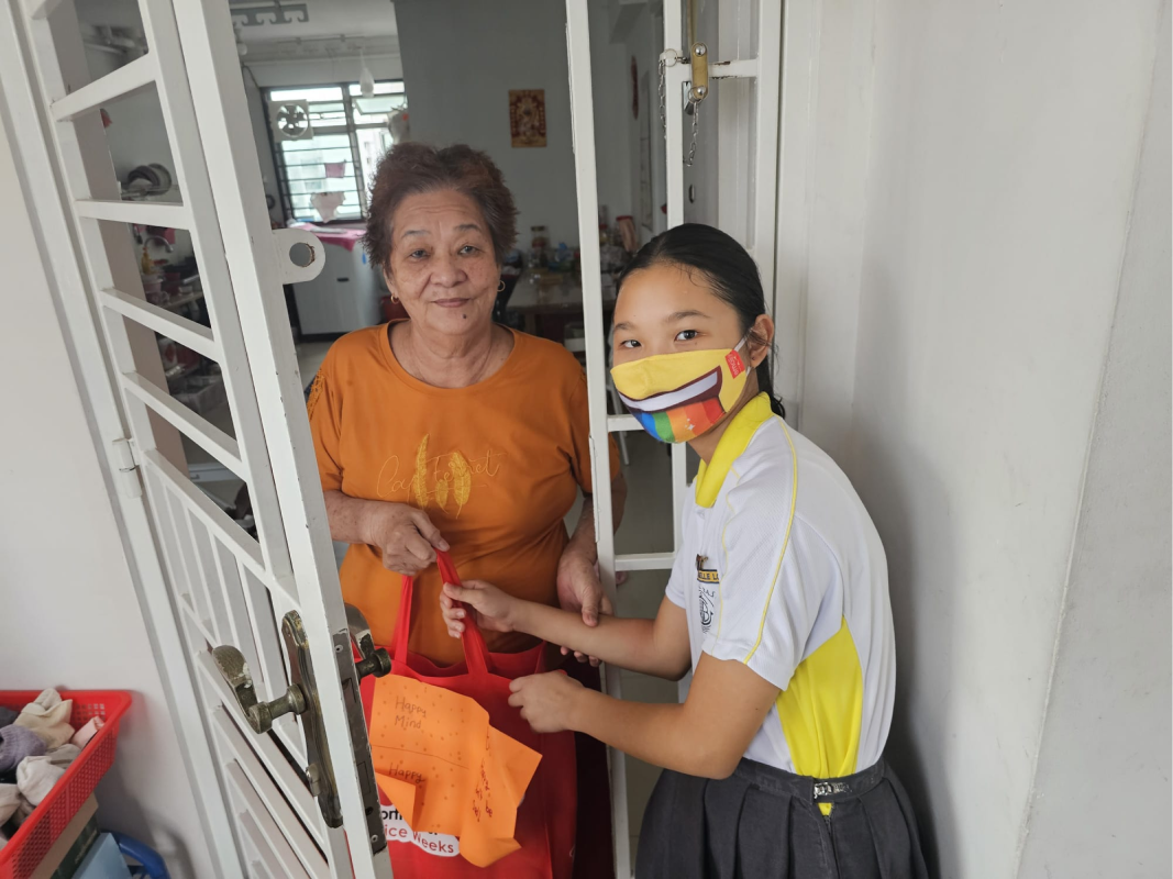 A masked schoolgirl in uniform hands a red bag to an elderly woman standing at her apartment door.