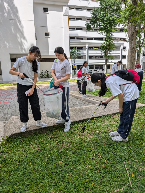 Students in white shirts pick up litter outdoors using grabbers and a plastic bag on a grassy area near a building.