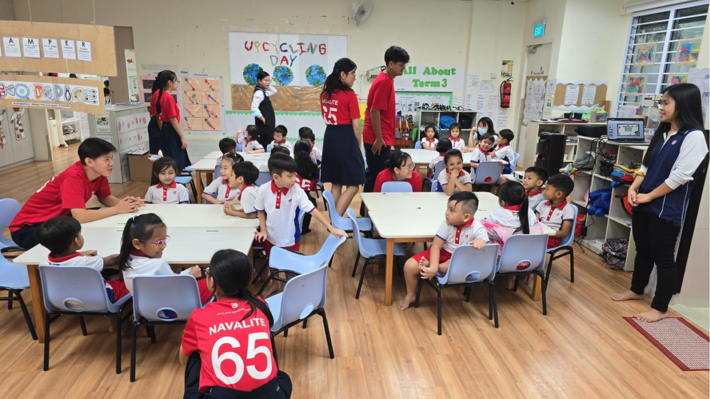 Young children sit at tables in a classroom decorated with an Upcycling Day banner, supervised by older students in red jerseys.