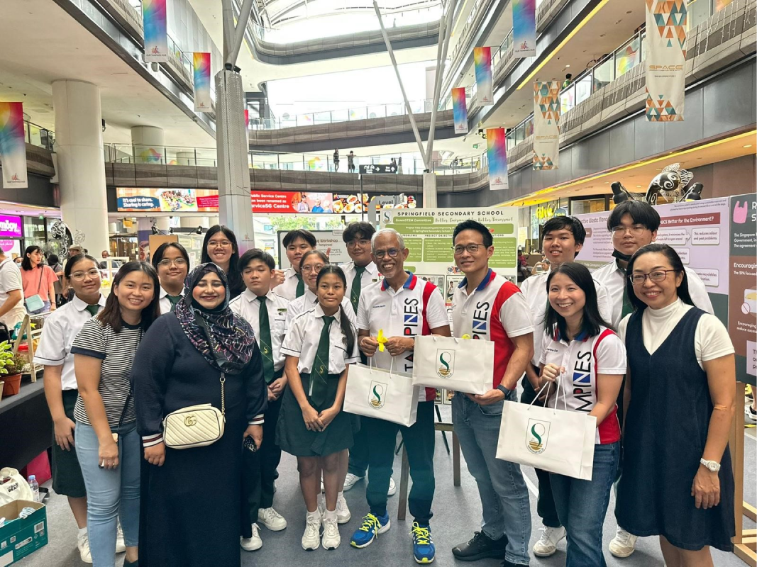 A group of students and adults pose together in a shopping mall atrium near a Springfield Secondary School display board.