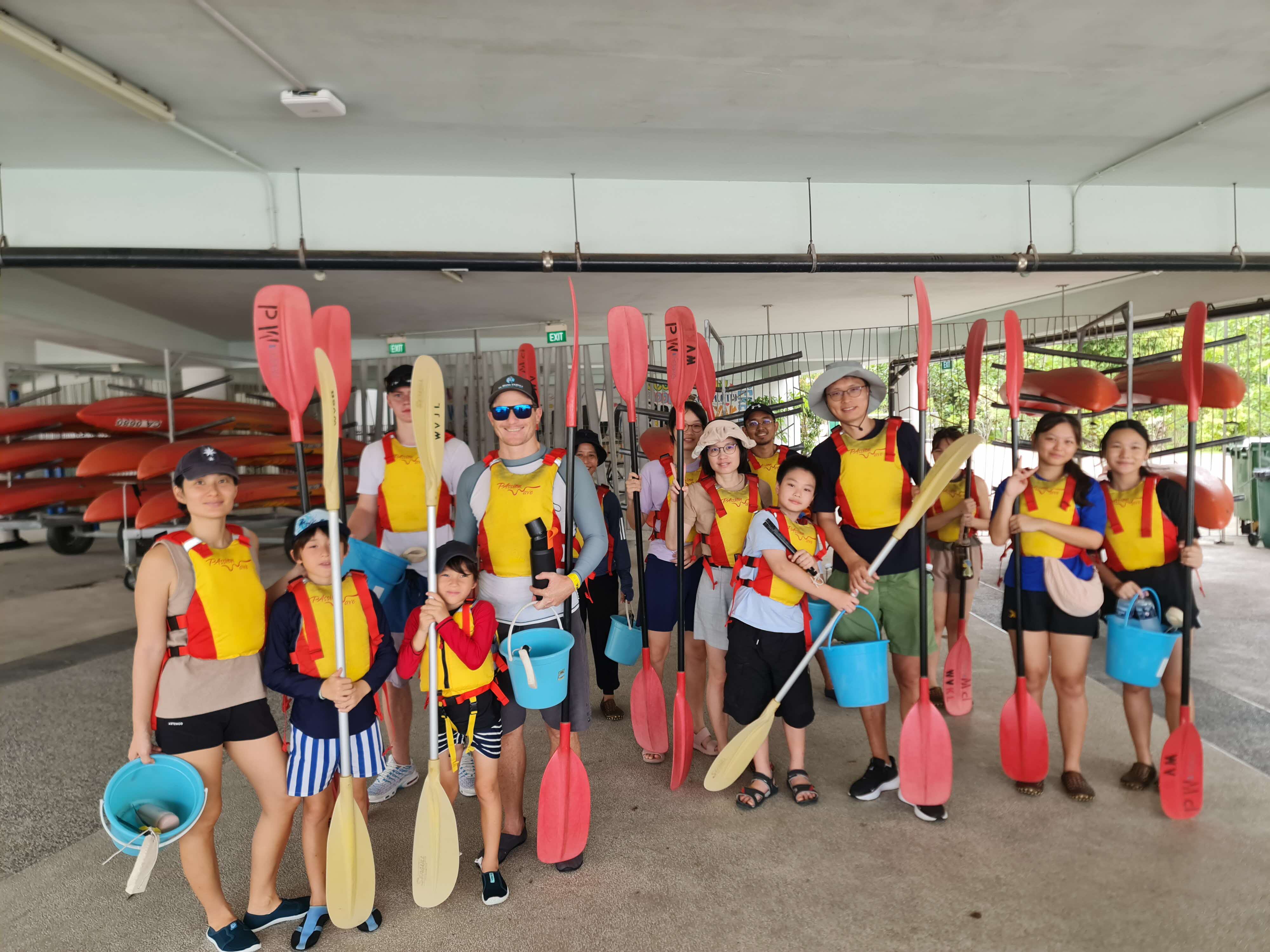 A group of adults and children in life vests hold paddles and buckets.