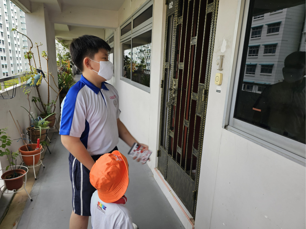 A masked boy in a school uniform and a small child in an orange hat stand outside an apartment door.
