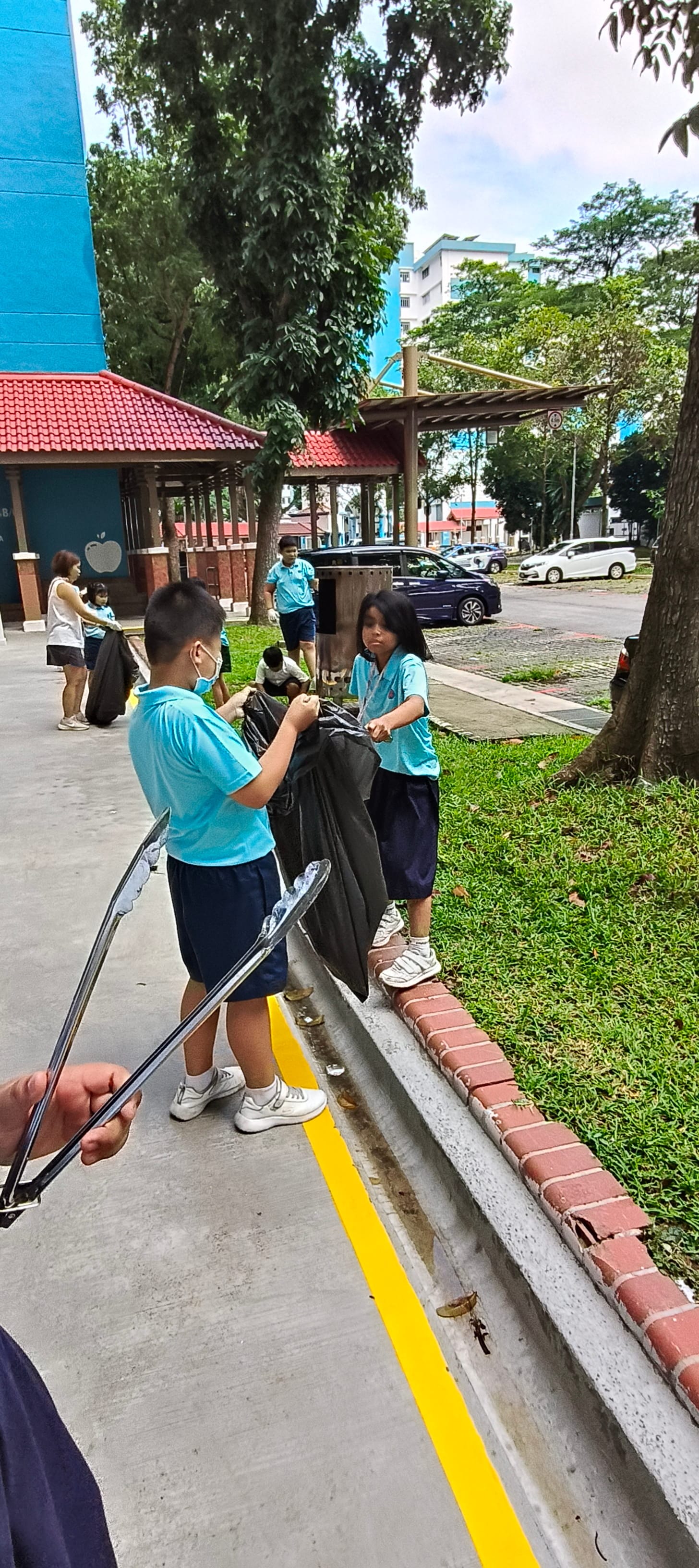Two children in blue uniforms collect trash in black bags outdoors near a school building with a red roof.