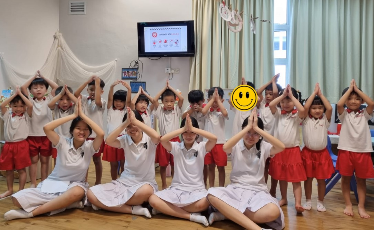 Young children and teachers in white and red uniforms pose with hands raised above their heads in a classroom.