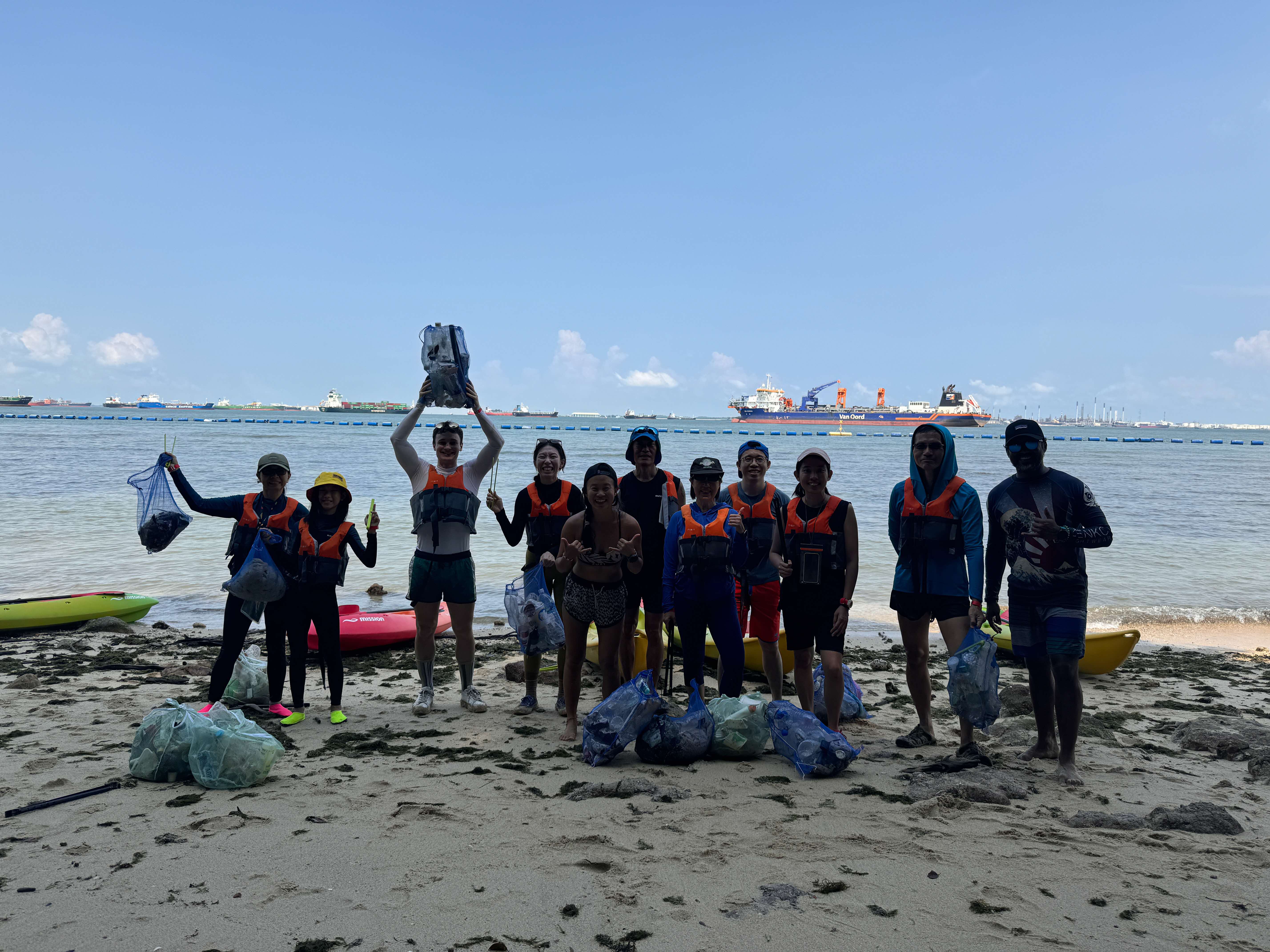 A group of people in life vests stand on a beach holding bags of collected trash, with ships visible on the water behind them.