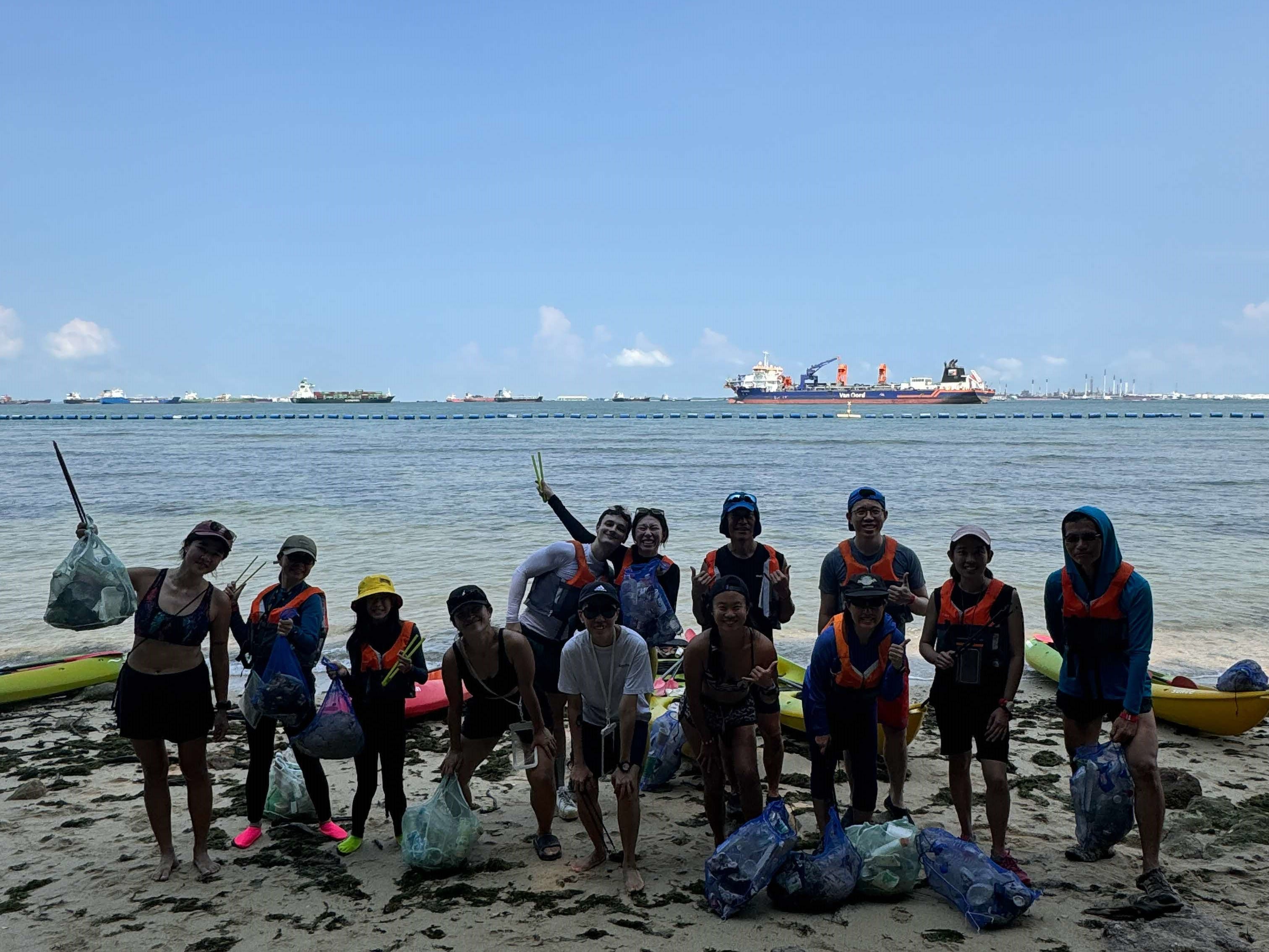 Group of people posing on a beach with collected trash bags and kayaks, ships visible on the water behind them.