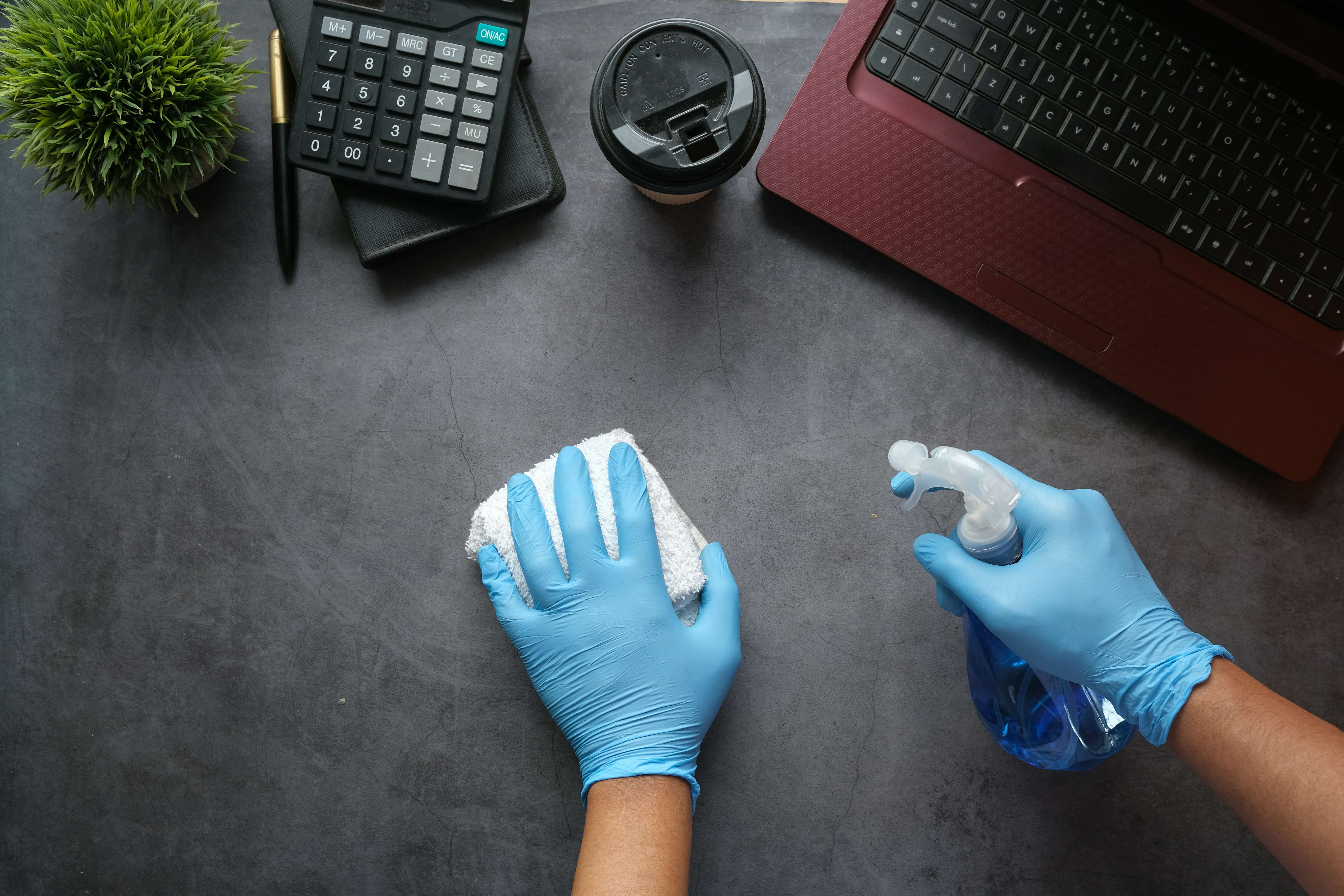 Gloved hands wipe a desk with a cloth and spray bottle near a laptop, calculator, coffee cup, and small plant.