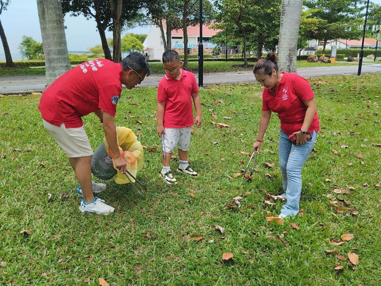 Three people in red shirts pick up litter with tongs on a grassy park area near a waterfront.