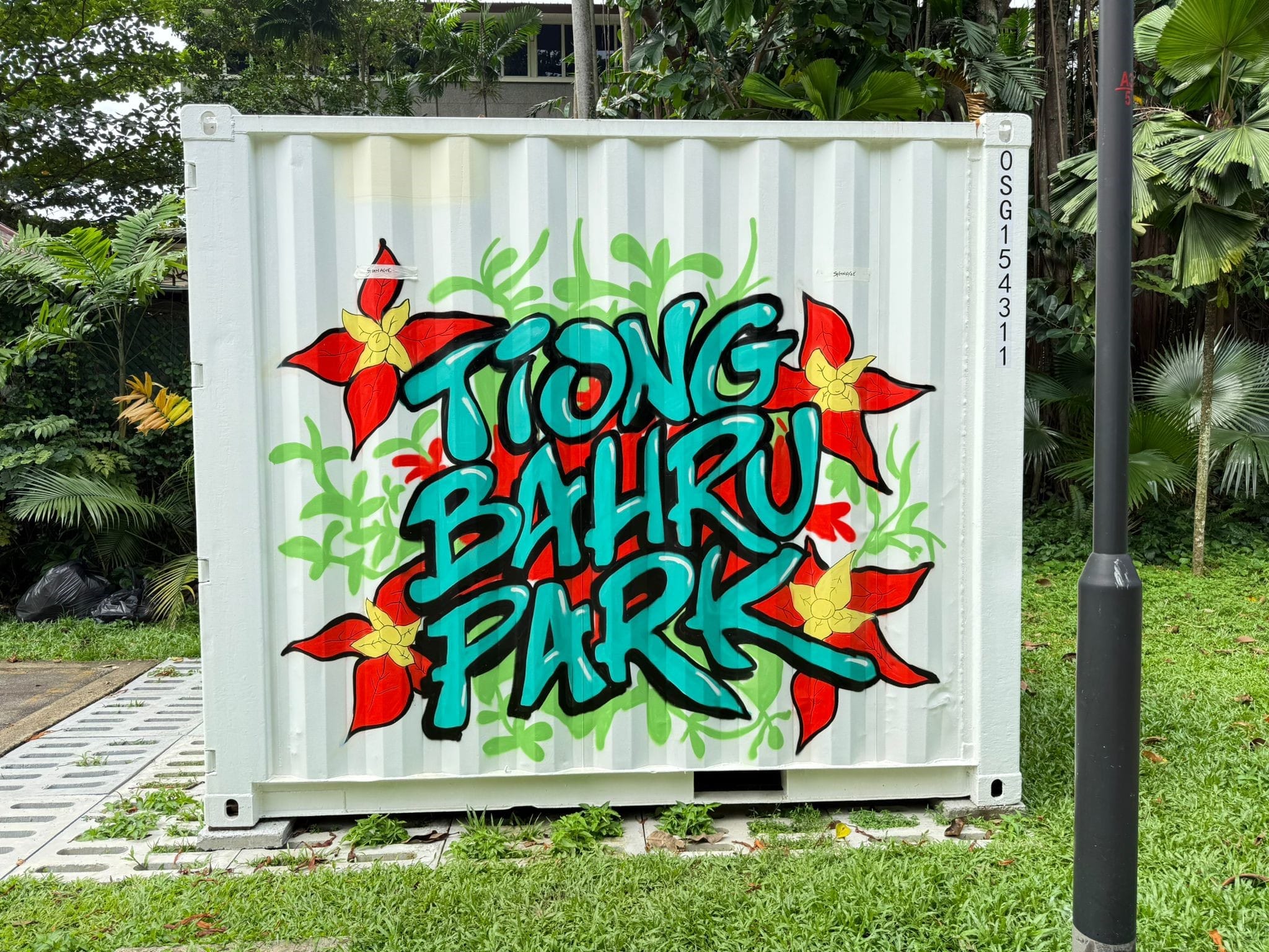 A white shipping container at Tiong Bahru Park, decorated with a vibrant graffiti-style mural featuring the words 'Tiong Bahru Park' in teal lettering, surrounded by red tropical flowers and green foliage.