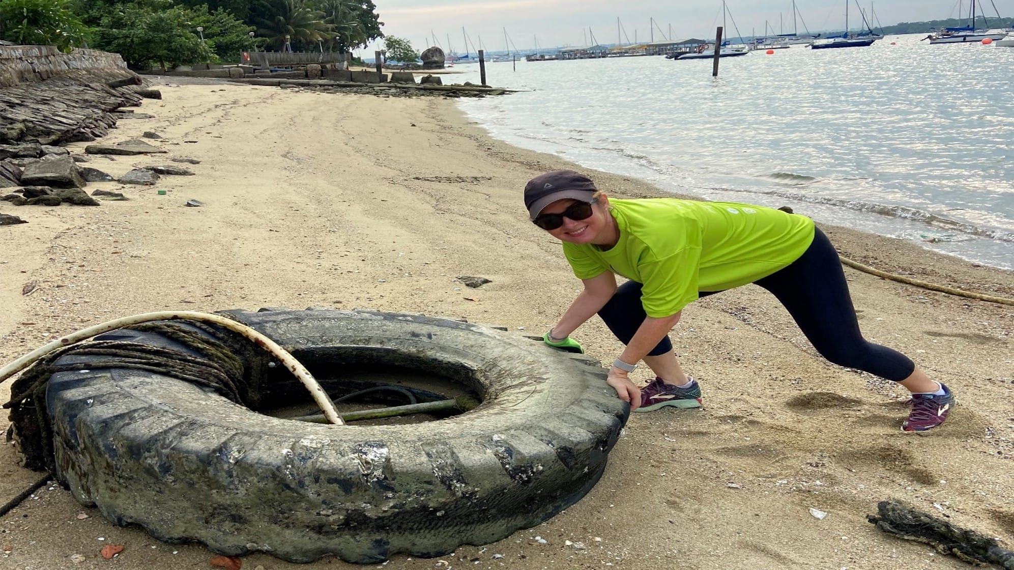 Woman in green shirt and cap pushes a large worn tire on a sandy beach with calm water and boats behind her.