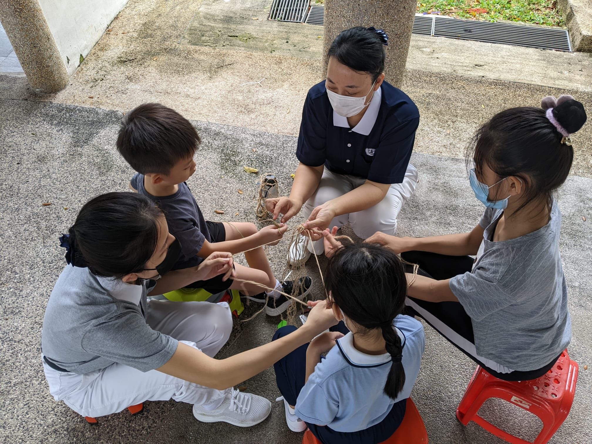 An adult wearing a mask guides three children in a rope-weaving activity while seated outdoors on a paved ground. Hai Yan educating neighbourhood residents on making reusable bags for Tzu Chi Environmental Sustainability Day