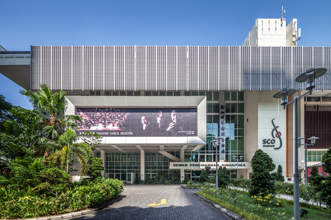Modern building with "Dewan Persidangan Singapura" sign, large billboard, greenery, and Singapore Chinese Orchestra logo.