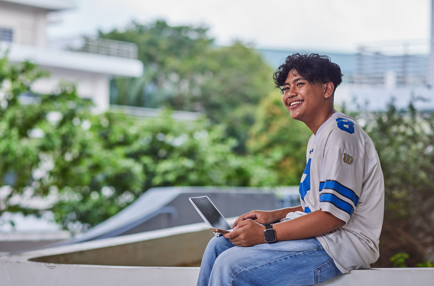 Student using a tablet outdoors on campus with greenery and buildings in the background.