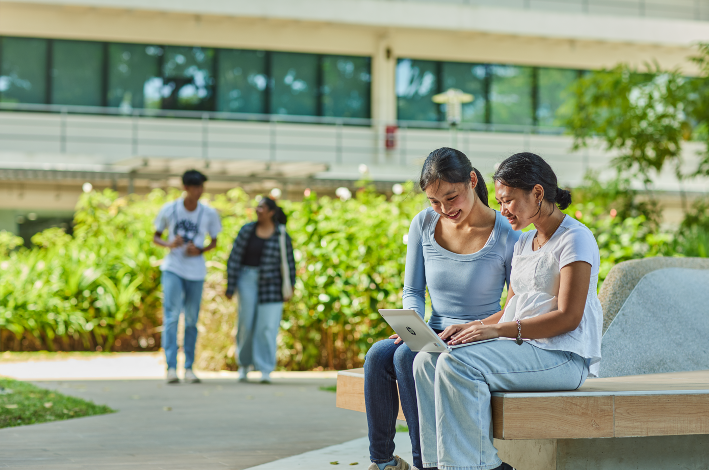 Two students sitting on a bench outdoors, looking at a laptop, with others walking in the background.