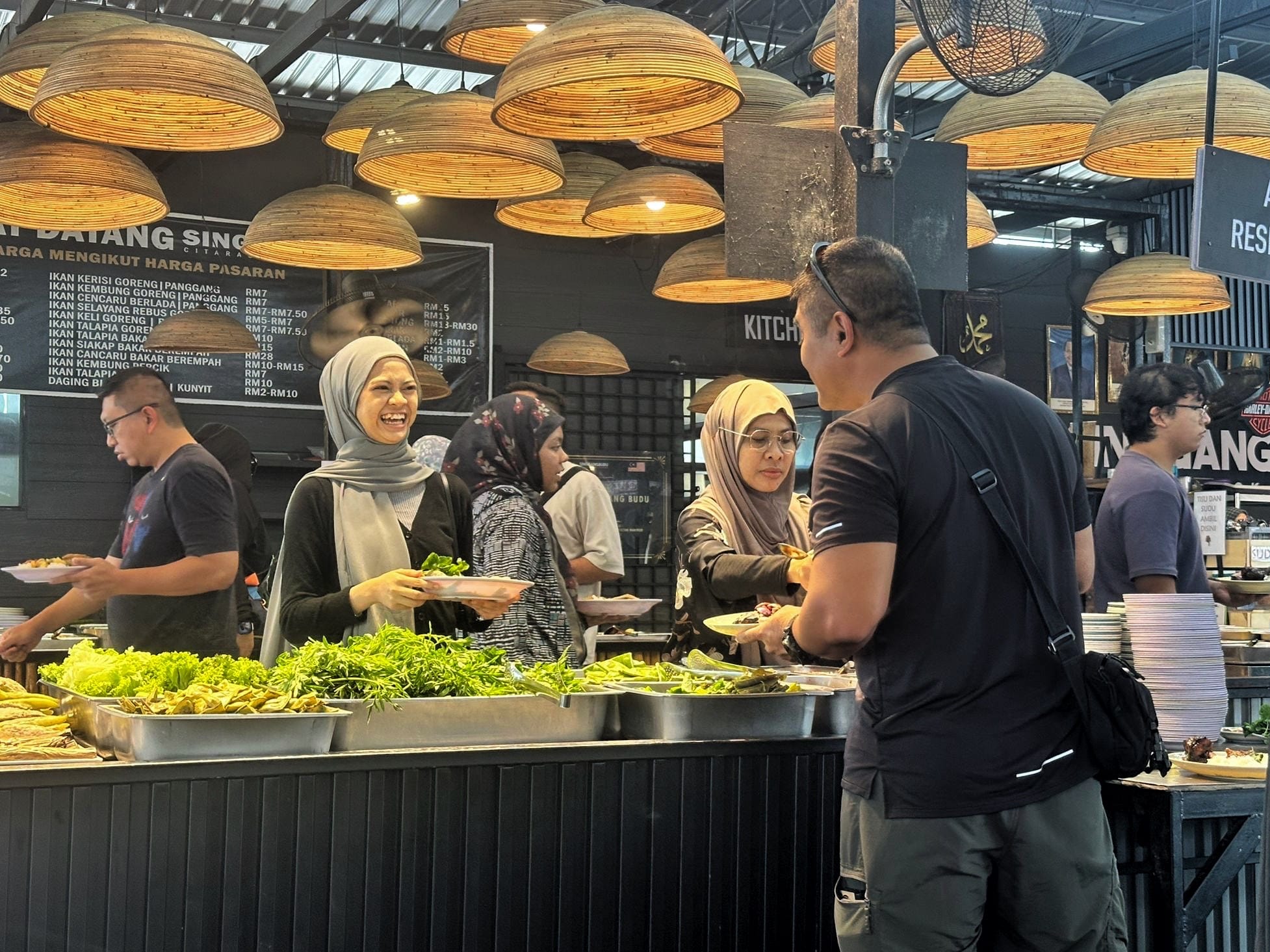 Group members selecting food from a buffet counter at a restauranyt, with trays of vegetables and dishes displayed beneath hanging woven lamps.