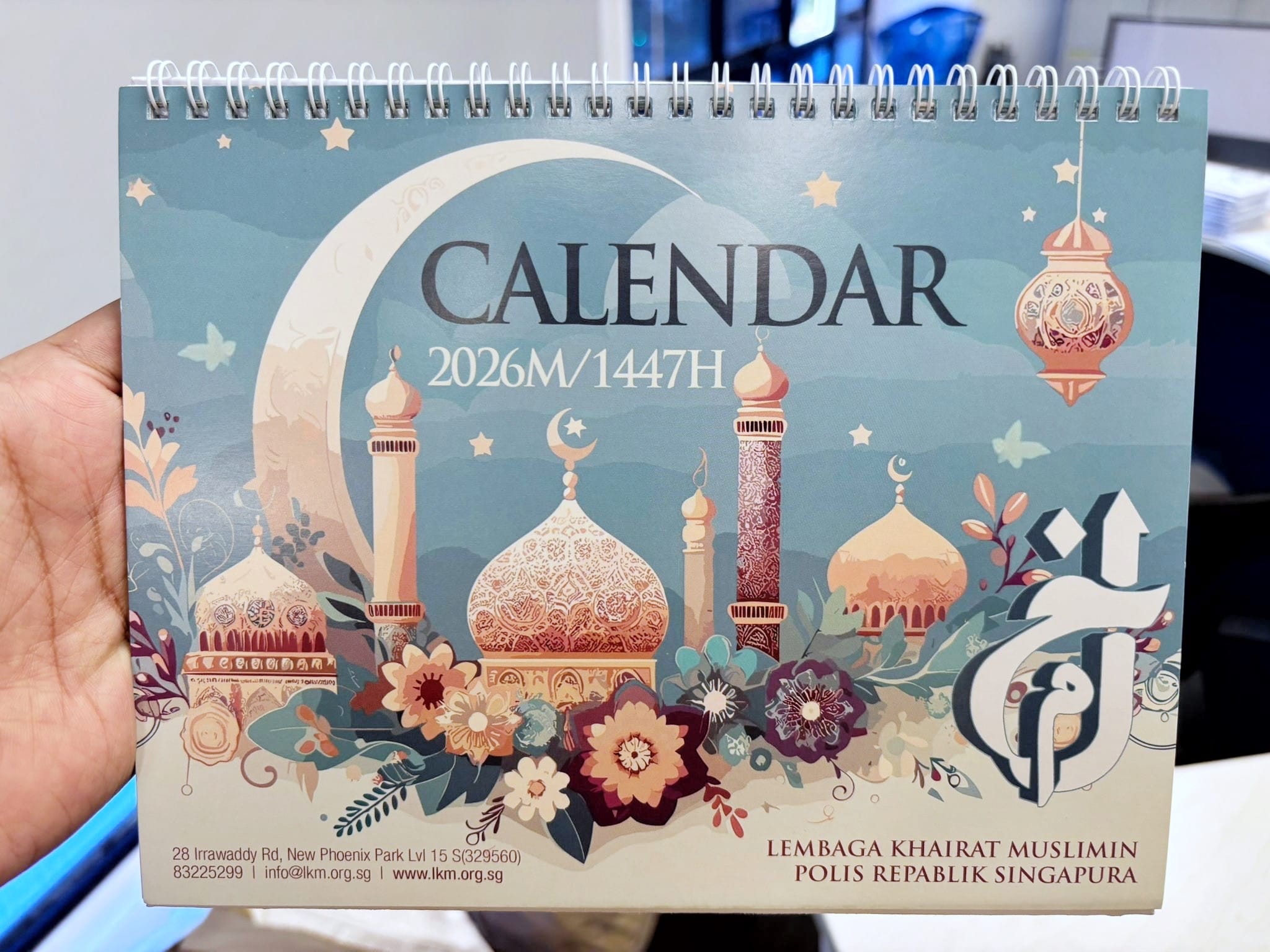 Hand holds spiral desk calendar showing mosque domes, crescent moon, and text CALENDAR 2026M/1447H.