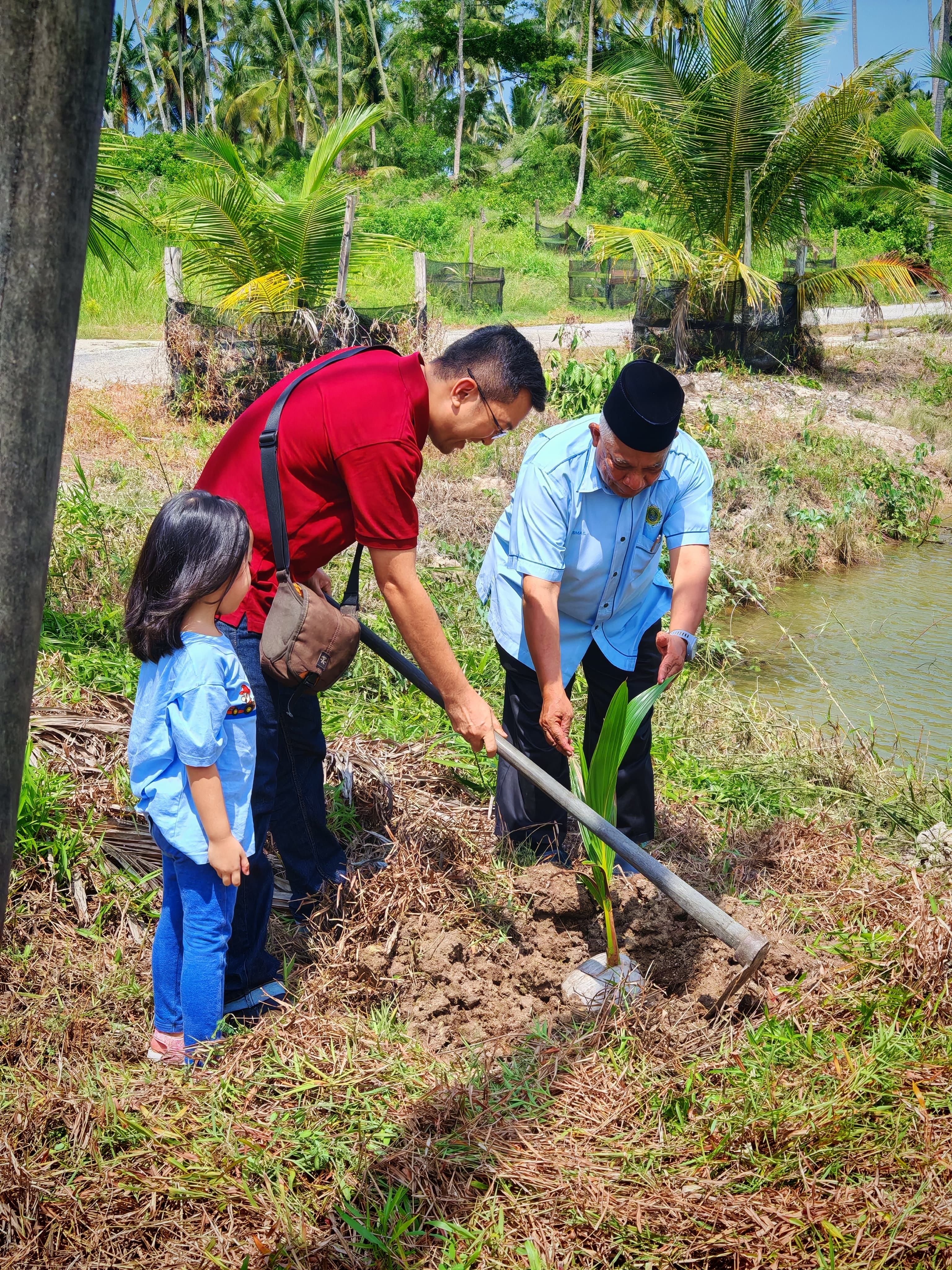 LKM Vice-President and an orphanage representative planting a young coconut tree beside a waterway while a child observes nearby.