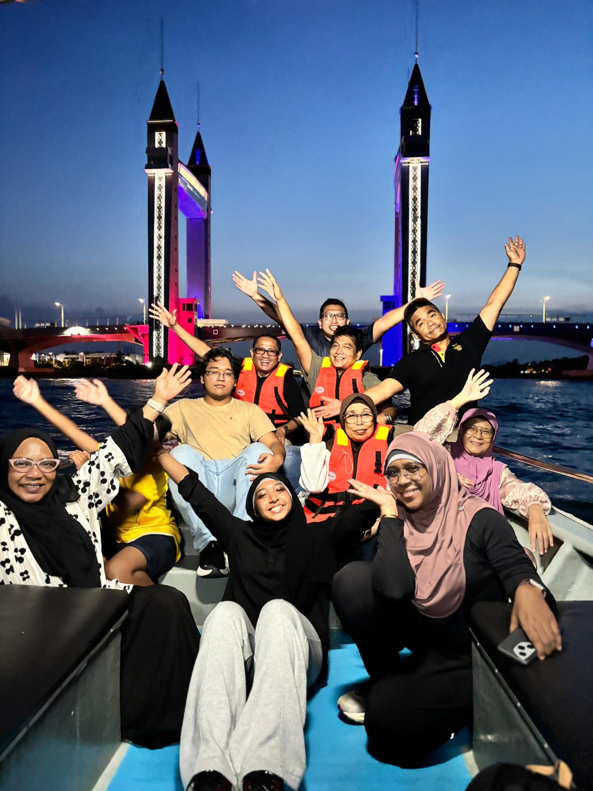 LKM members posing on a river cruise boat at dusk with an illuminated drawbridge in the background.