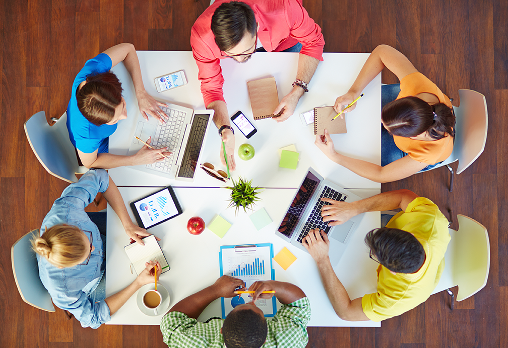 Overhead view of a diverse team collaborating around a table with laptops, tablets, charts, and notes, representing teamwork and project planning.