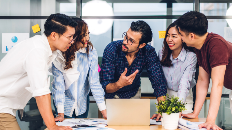 Five people around a table with a laptop and papers in an office.