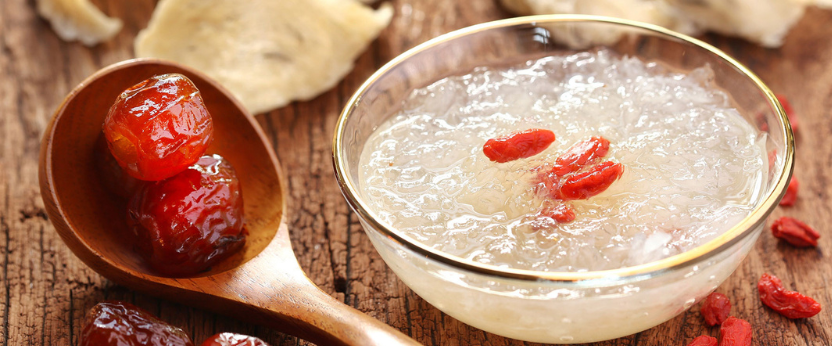 A clear glass bowl filled with a translucent, gelatinous soup sits next to a wooden spoon holding red dates.