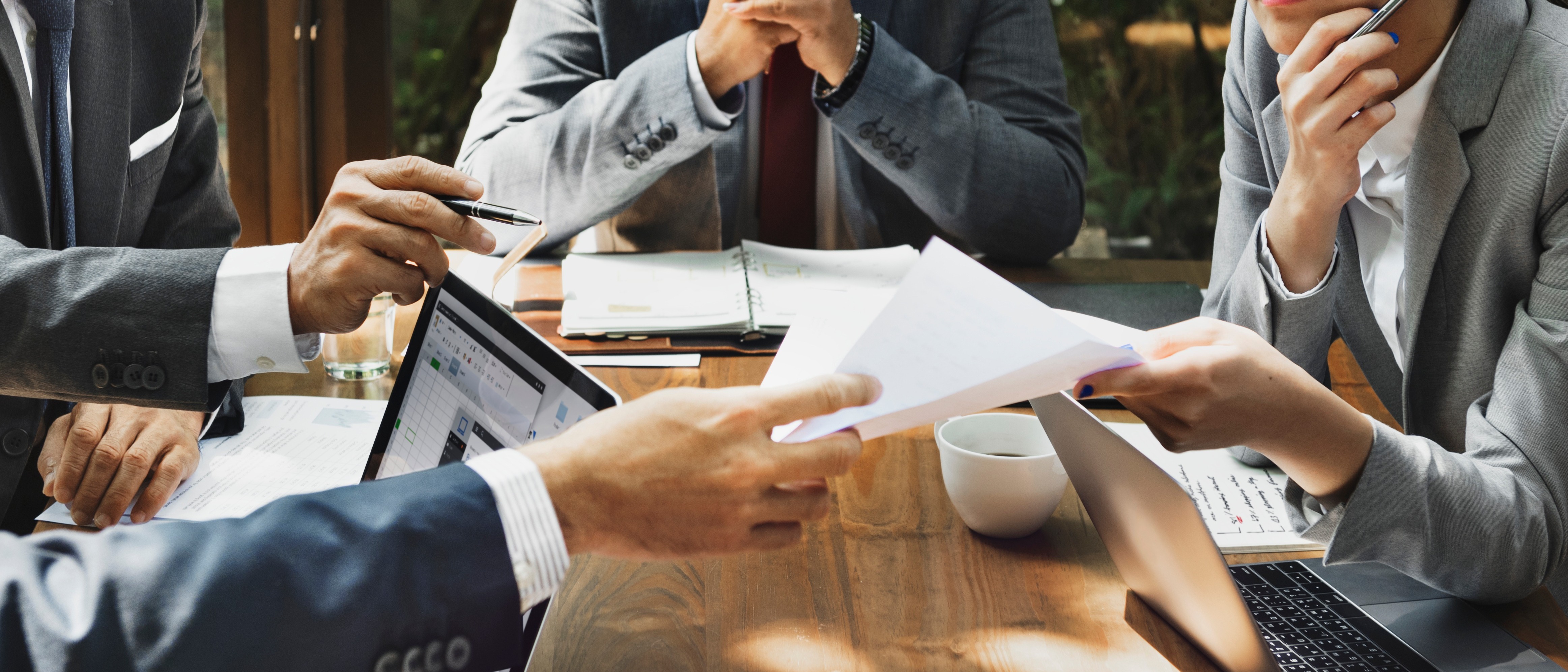 Four professionals in suits discuss documents around a wooden table.