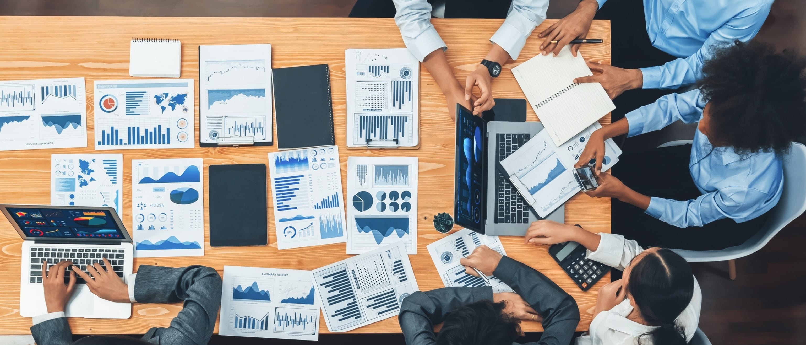 A diverse group in professional attire collaborates at a table cluttered with charts, graphs, and laptops. 