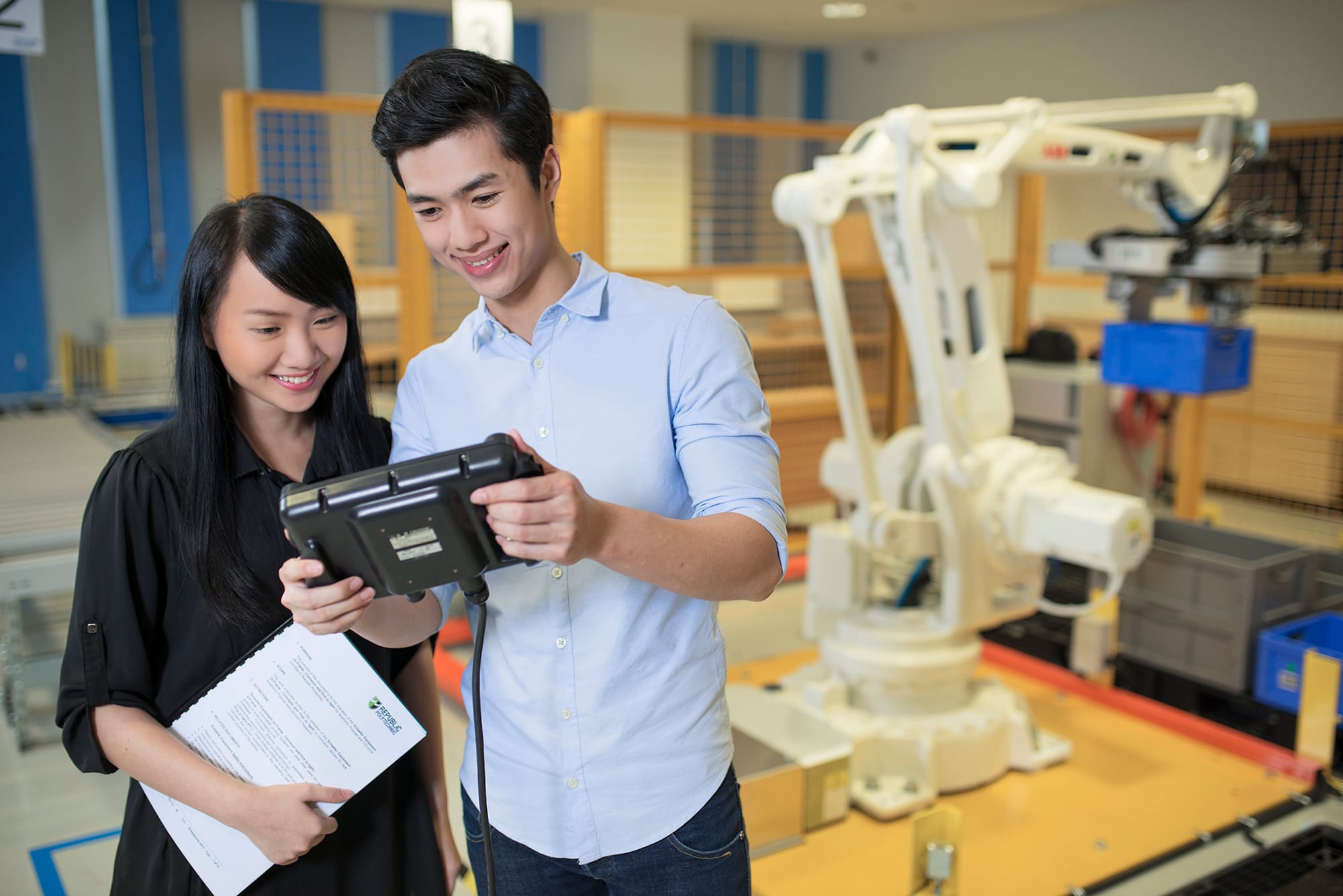 Two people standing together in a robotics lab, smiling at a tablet. 