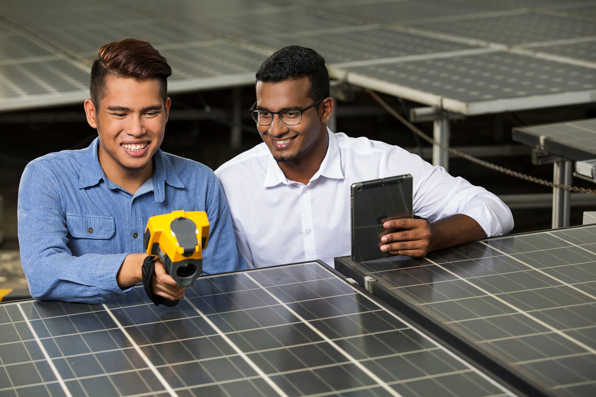 Two men examining solar panels; one uses a thermal camera, the other holds a tablet.