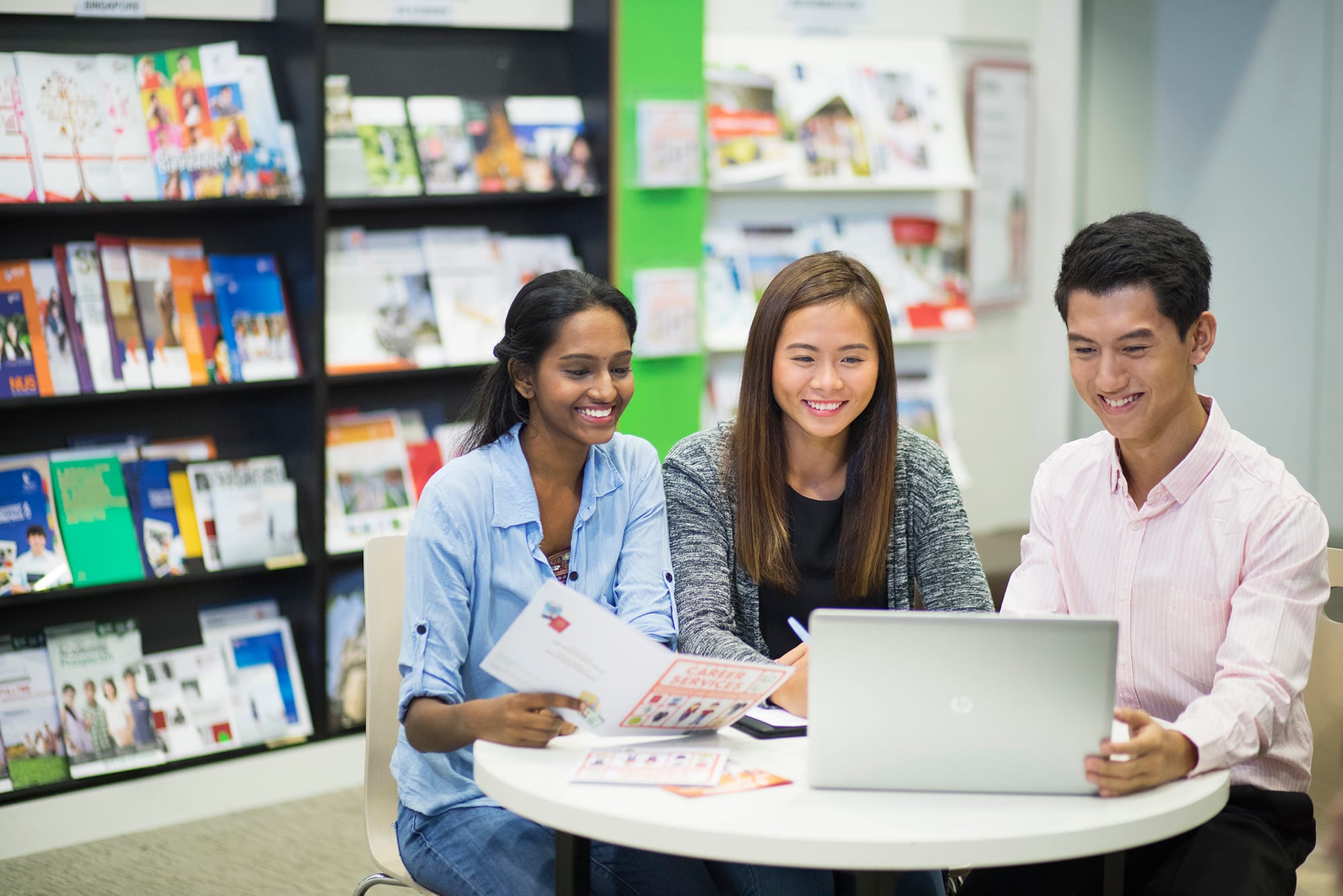 Three students sit around a table, smiling and collaborating on a laptop in a library.