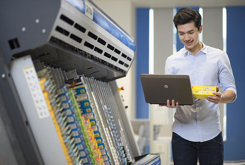 Man using a laptop to check packaged products beside a printing or production machine.