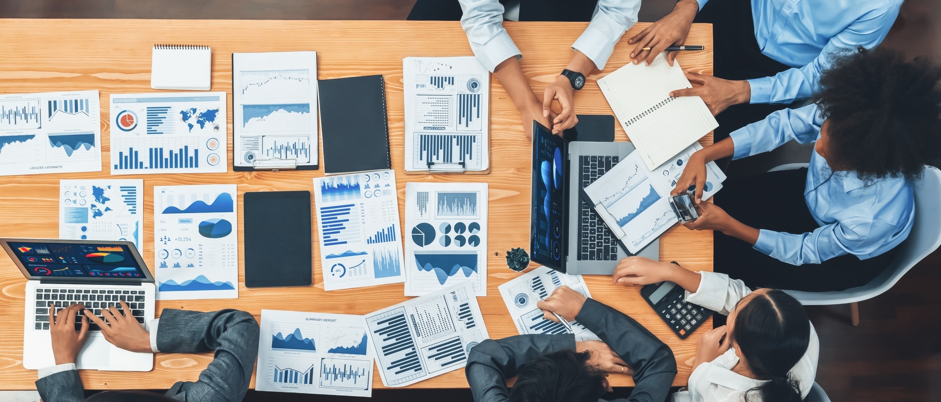 Team collaborating around a table covered with financial reports, dashboards, and laptops, representing business intelligence and data-driven decision-making.