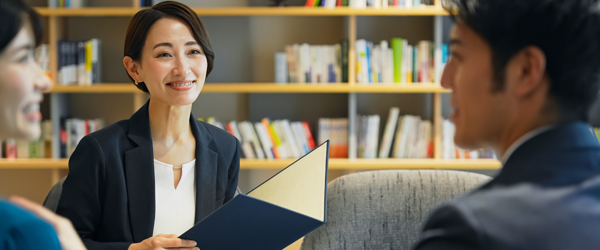 Three people in suits sit with open blue folder, bookshelf in background.