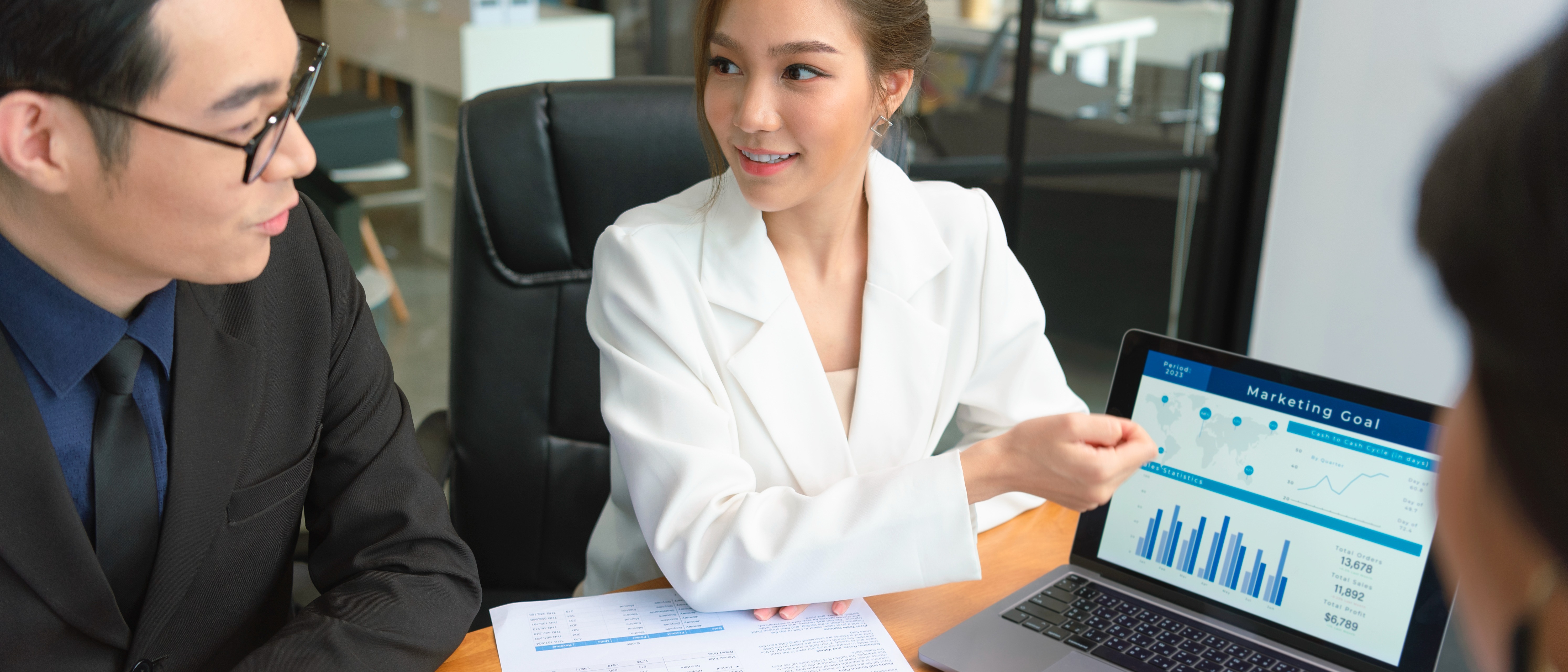 A woman in a white blazer presents a marketing graph on a laptop to two colleagues in a meeting room.