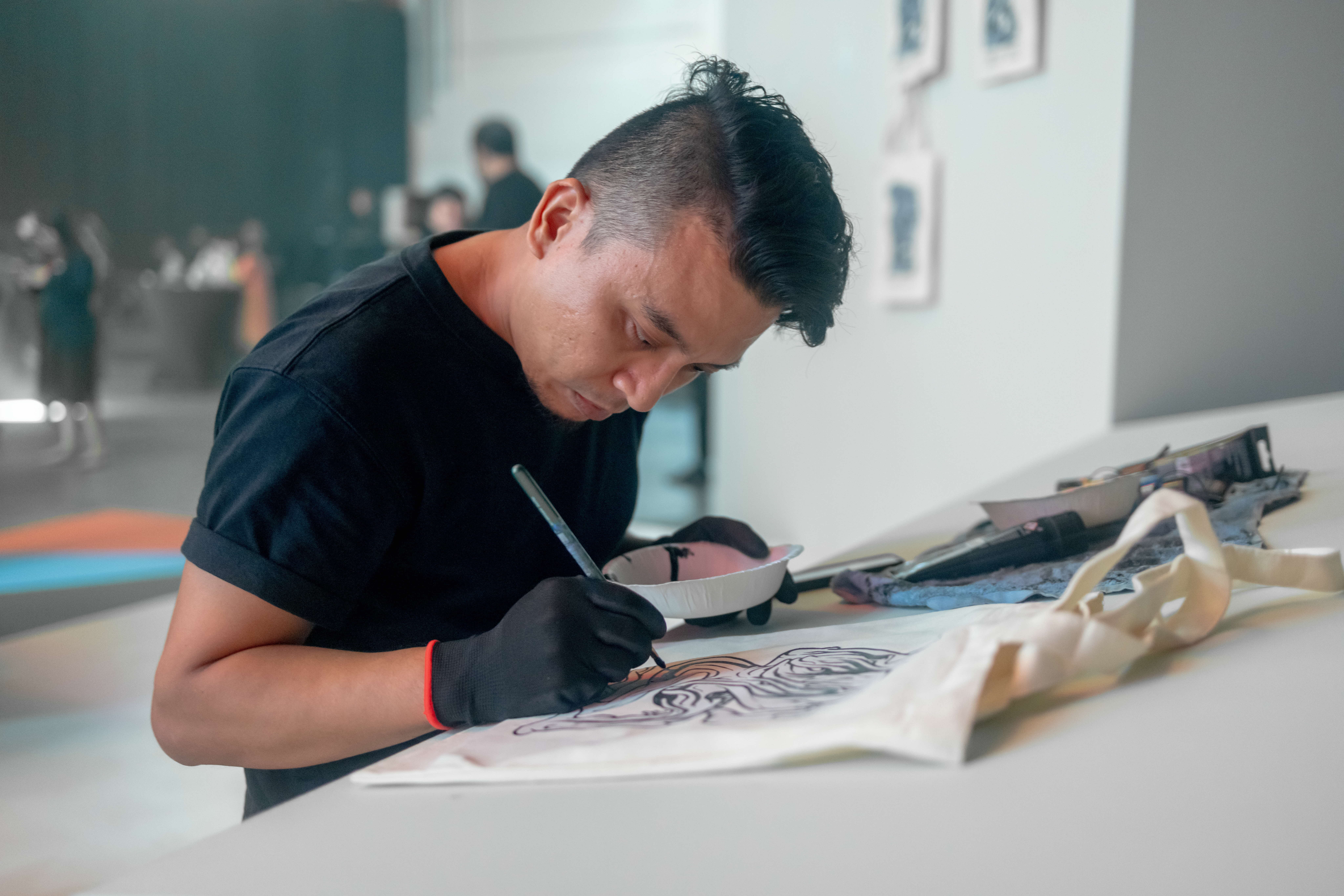 Man in black T-shirt and gloves leans over a table, drawing on paper with a brush and ink tray nearby.