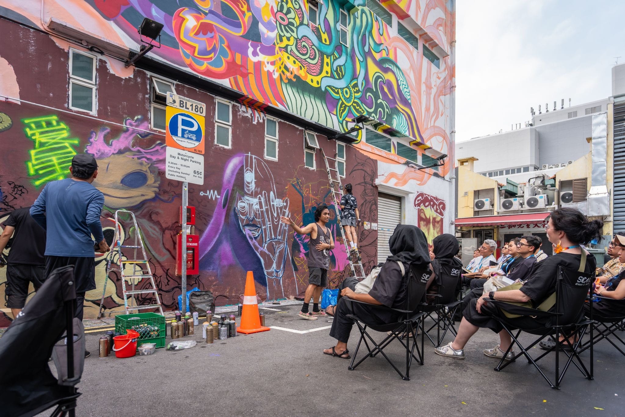 Artist paints graffiti mural on a wall while seated spectators watch; ladders, spray cans, and a cone nearby.