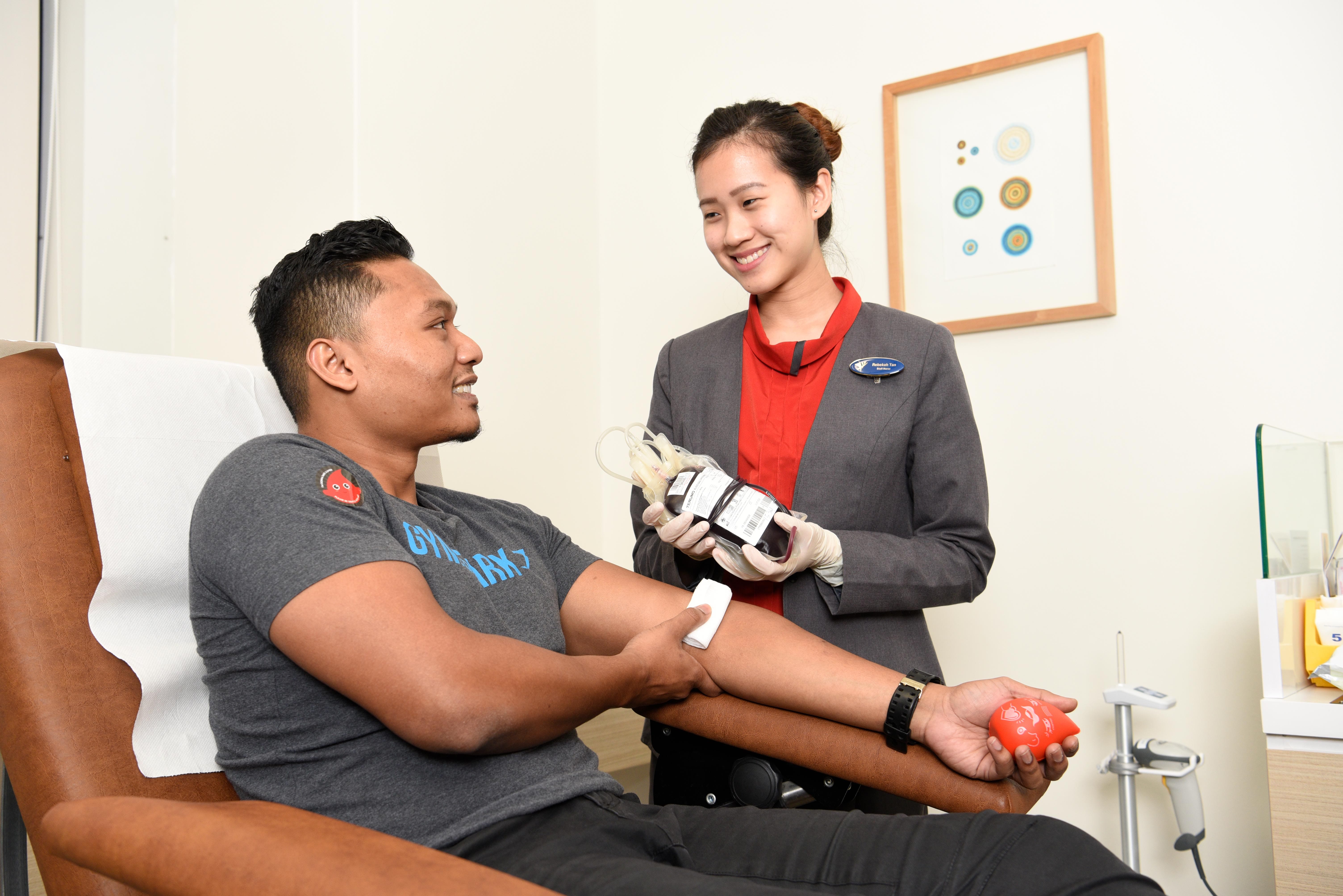 Nurse holds a blood bag while smiling at a seated donor squeezing a heart ball in a clinic.