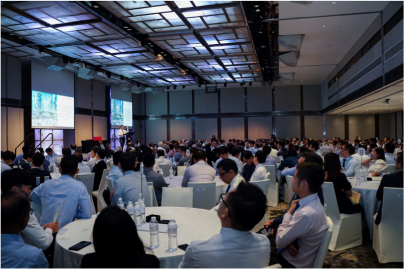 A large conference hall filled with attendees seated at round tables, facing a speaker at a podium with projection screens.