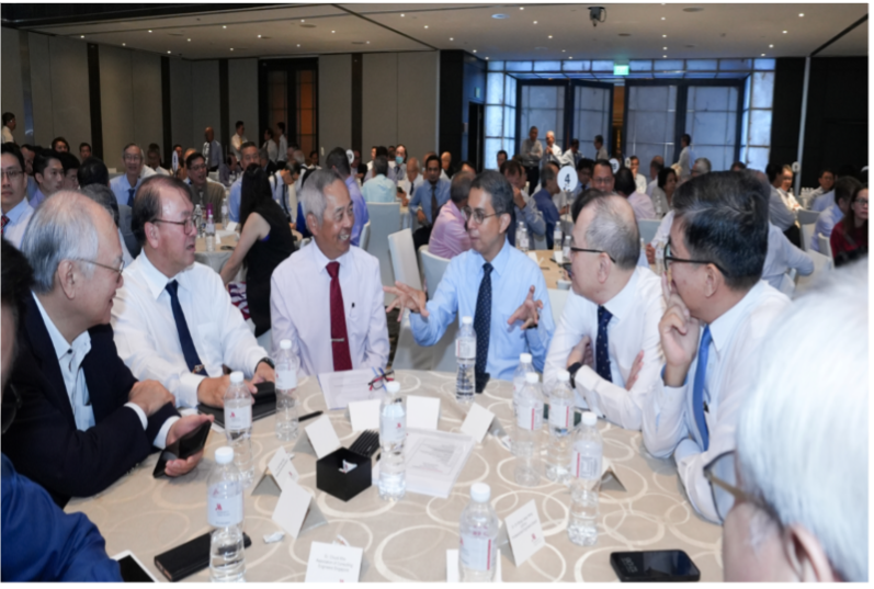 Several men in dress shirts and ties converse around a round table in a large, crowded conference banquet hall.