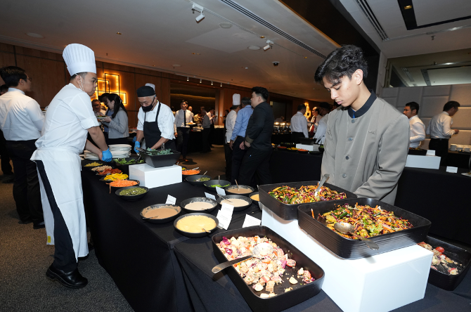 Chefs and staff in uniforms tend to a large buffet spread with multiple dishes in a banquet hall.