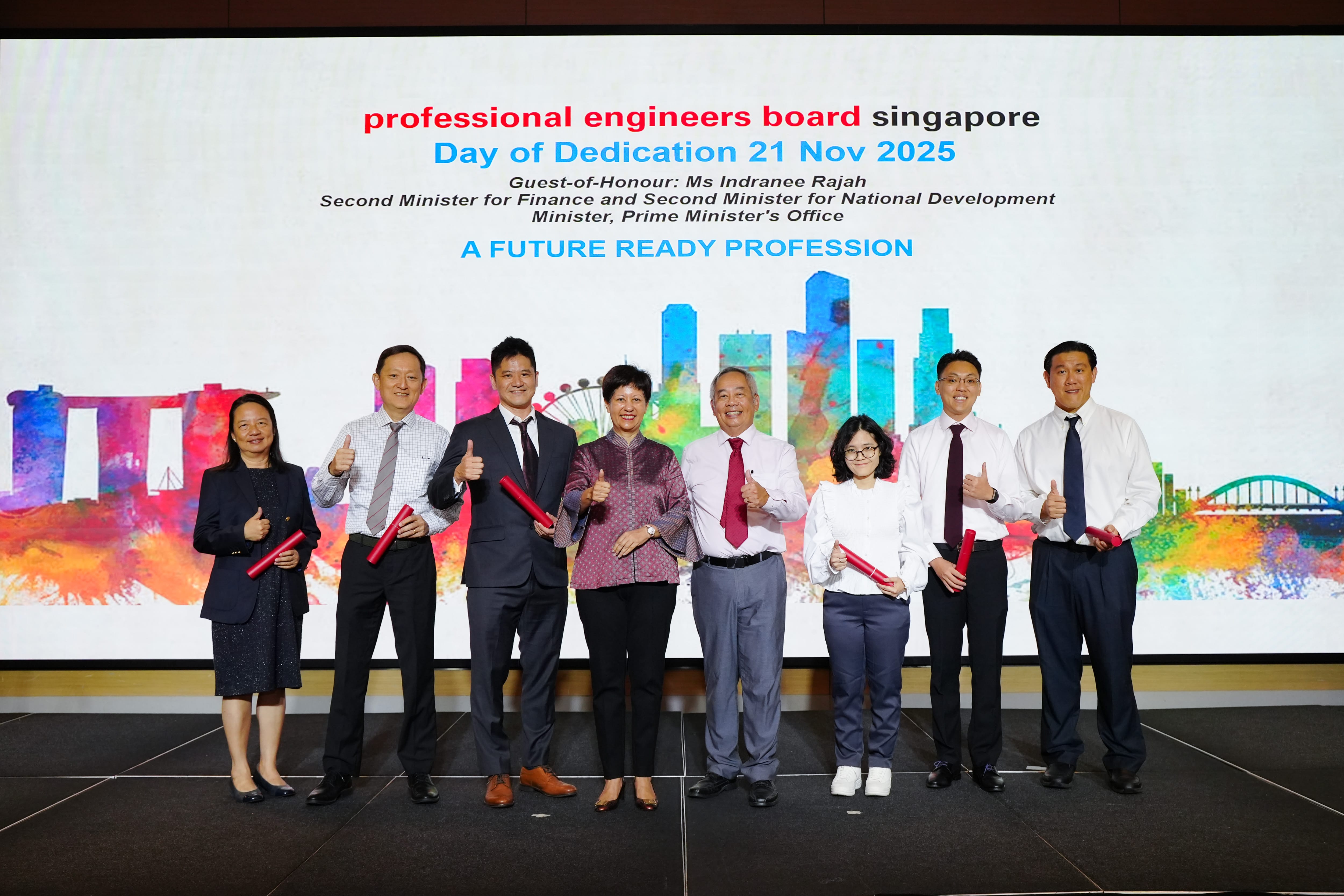 Eight people pose with thumbs up holding red scrolls at the Professional Engineers Board Singapore Day of Dedication 2025.