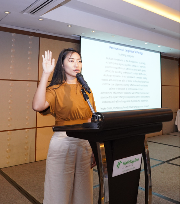 Woman in orange top stands at a Holiday Inn podium, raising her right hand while reciting the Professional Engineer's Pledge.