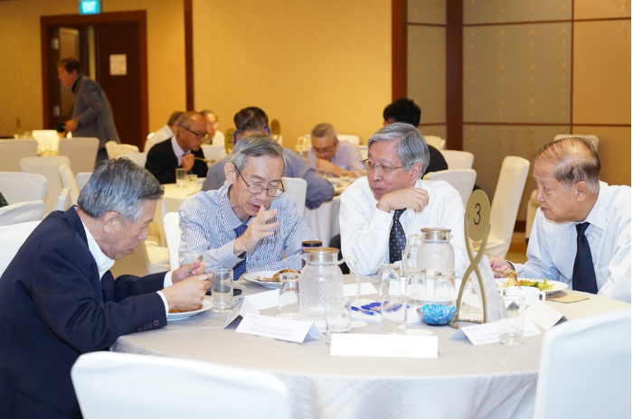 Four older men in formal attire sit around a round table in a banquet hall, engaged in conversation.