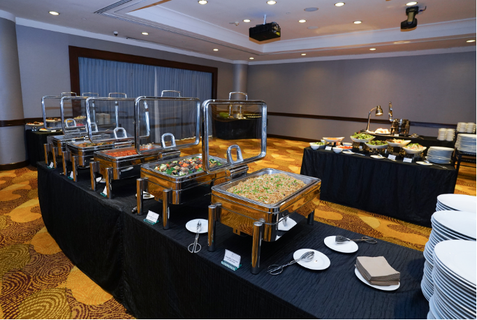 A catered buffet setup with multiple chafing dishes of hot food, stacked white plates, and a salad station in a banquet hall.