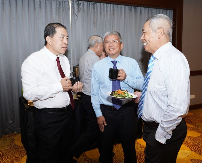 Three men in dress shirts and ties chat at a reception, one holding a mug and plate of salad.