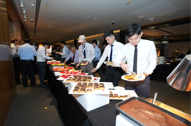 People in formal attire serving themselves at a long buffet table with various dishes and pastries.